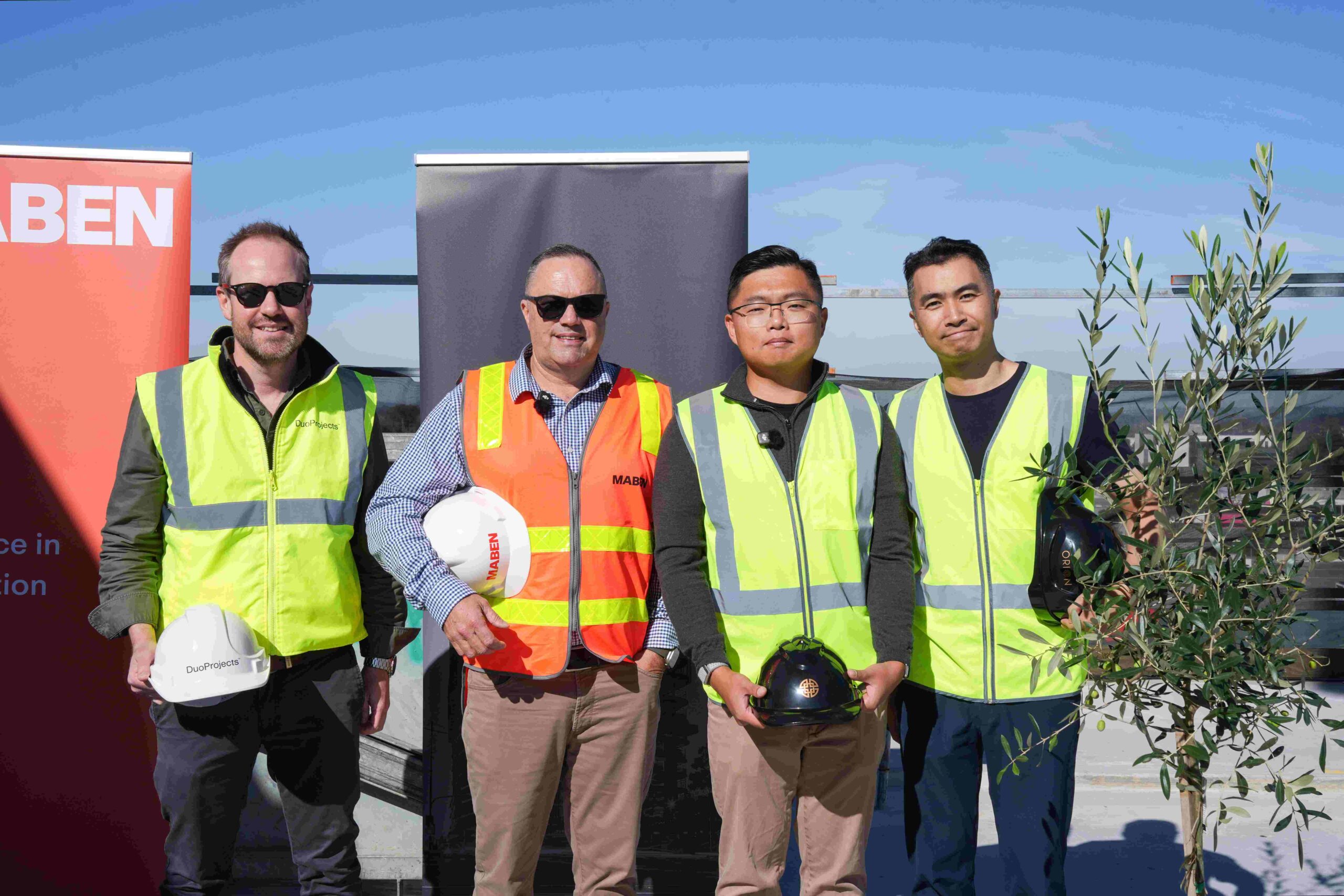 Four males in high vis vests holding hard hats pose for the camera on building rooftop, all holding hard hats and standing next to a potted tree as part of a topping out ceremony at a retirement apartment development in Ringwood